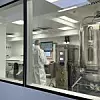Blick durch ein Fenster in eine Reinraum-Produktionseinrichtung; eine Person im weißen Reinraum-Overall steht vor den Bedienelementen eines Bioreaktors.