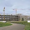 Im Bau befindliches Firmengebäude auf das eine Straße zugeht umrandet von grün mit Baukränen im Vordergrund
