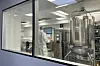 Blick durch ein Fenster in eine Reinraum-Produktionseinrichtung; eine Person im weißen Reinraum-Overall steht vor den Bedienelementen eines Bioreaktors.