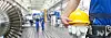 Arbeiter mit Helm und Handschuhen in der Hand im Vordergrund, im Hintergrund Werkshalle mit weiteren Arbeitern