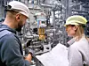 Zwei Beschäftigte mit Schutzhelmen prüfen Pläne vor einer Industrieanlage mit Rohren.