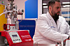 Laborgerät mit rotem Bedienfeld neben einem Wissenschaftler in weißem Kittel.