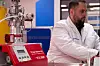Laborgerät mit rotem Bedienfeld neben einem Wissenschaftler in weißem Kittel.