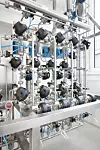 Komplexe industrielle Rohrleitungen mit Ventilen, Sensoren und Metallgestell in einem hellen Technikraum.