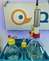 Versuchsaufbau mit mehreren Flaschen, Schläuchen und einem Metallzylinder vor einem Logo im Labor.