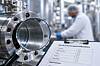 Industrieanlage mit Metallrohr und Prüfprotokoll im Vordergrund, Mitarbeiter unscharf im Hintergrund.