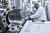 Industrieanlage mit Metallrohr und Prüfprotokoll im Vordergrund, Mitarbeiter unscharf im Hintergrund.