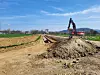 Bagger neben einem Erdwall an einer offenen Baugrube auf einem Feldweg in ländlicher Umgebung.
