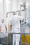 Zwei Arbeiter in weißer Schutzkleidung an einer industriellen Anlage mit Schläuchen und Metallbehältern.