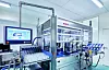 Automatisierte Industrieanlage mit Bildschirm, Förderband und Maschinen in einem hellen Produktionsraum.