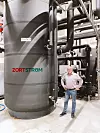 Mann steht in einer Industriehalle neben einem großen grauen Tank und mehreren Rohrleitungen.